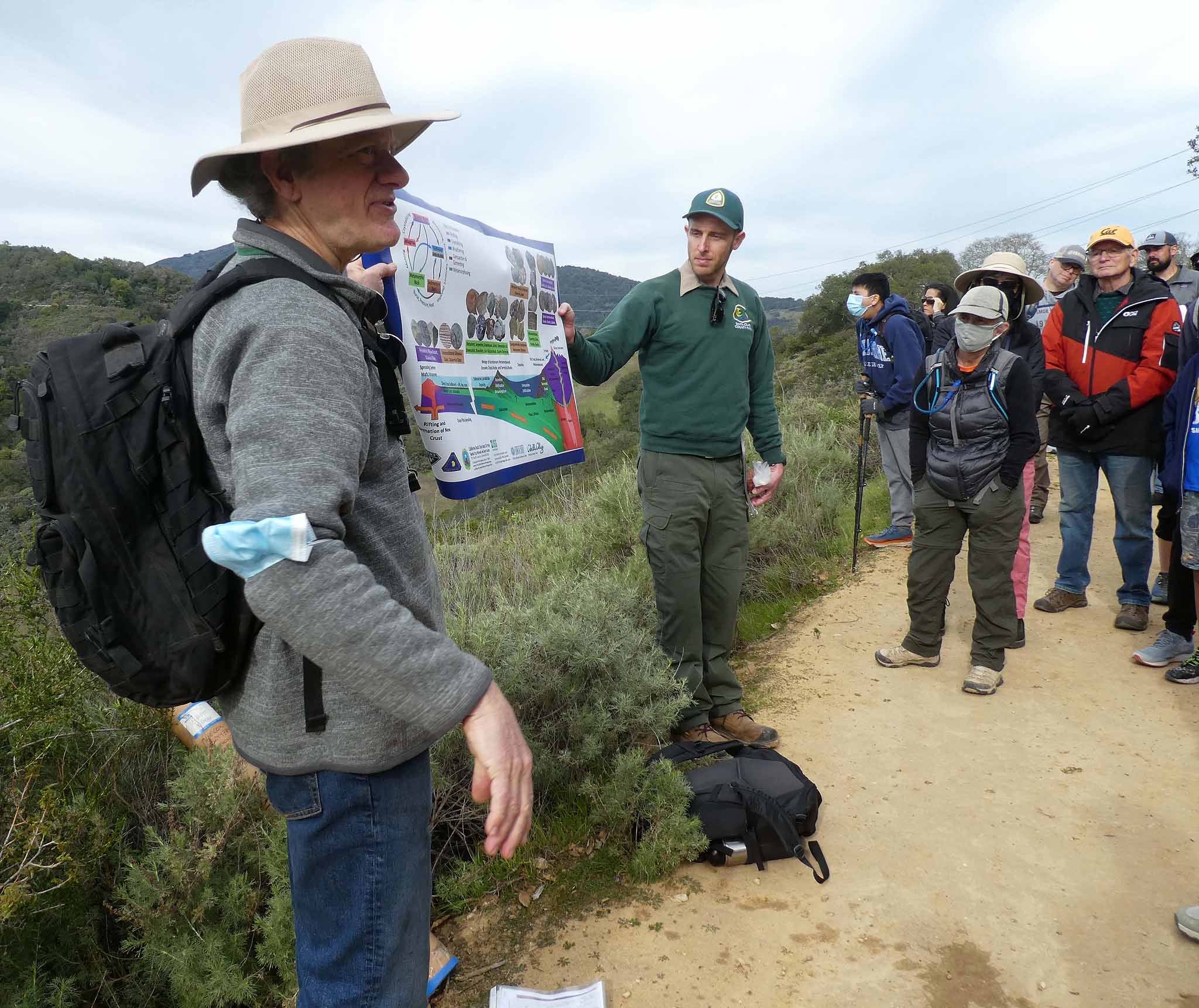 Michael Cox holds a poster about the rock cycle along with a Santa Clara County Park Interpreter while talking with the public on a trail.