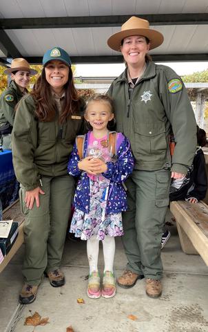 Child smiling, standing between two smiling santa Clara County Parks staff with third staff in background