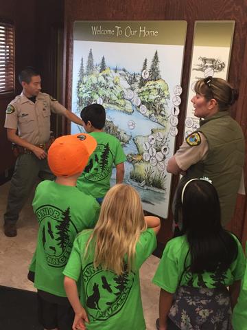 Ranger pointing to a "Welcome to our Home" habitat poster while Junior Rangers look at the poster